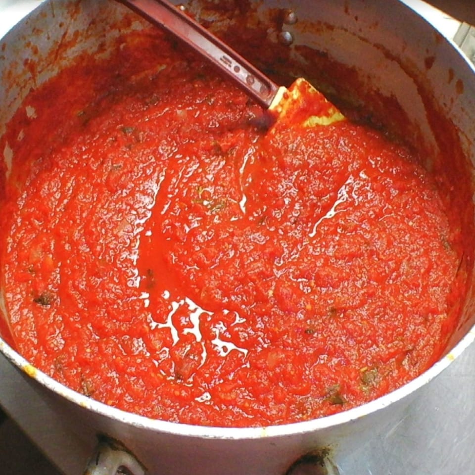 Large pot filled with Twisted Pies Pizzeria's rich red pizza sauce, with a spatula inside.